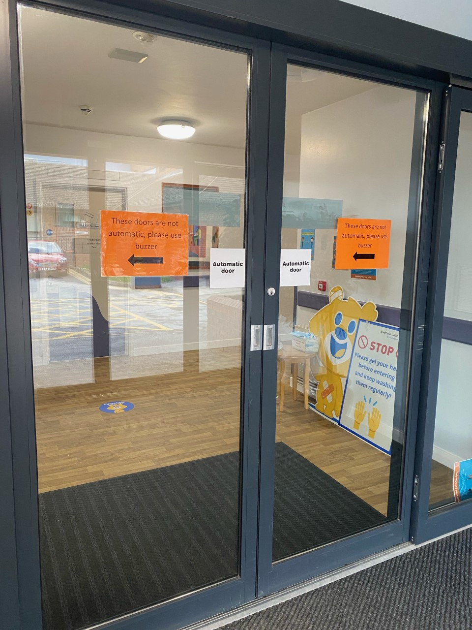 Photograph of set of doors with orange signs in windows saying 'These doors are automatic, please use buzzer' with an arrow pointing to the left