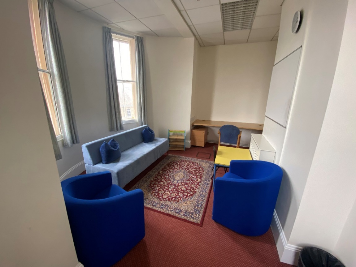 Photograph of a blue sofa and chairs with a rug and table. There is a desk and a whiteboard on the wall opposite the big windows