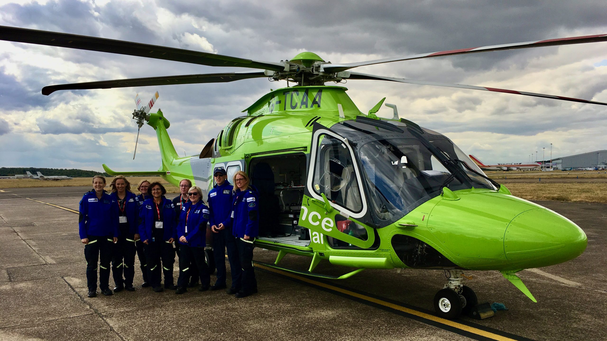 Photograph of staff wearing blue jumpers stood in front of a green helicopter, smiling